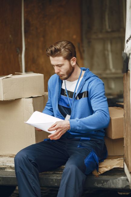 A man reviewing papers while sitting on the edge of a moving van in a storage area. He is dressed in a blue hoodie and dark work trousers, with a white lanyard around his neck. Behind him, there are s