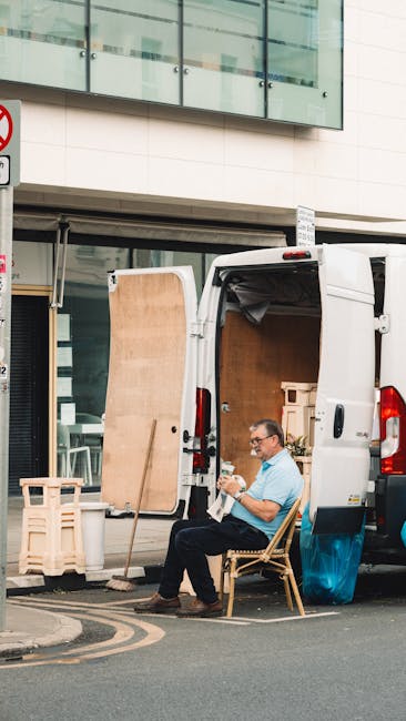 A man reviewing papers while sitting on the edge of a moving van in a storage area. He is dressed in a blue hoodie and dark work trousers, with a white lanyard around his neck. Behind him, there are s